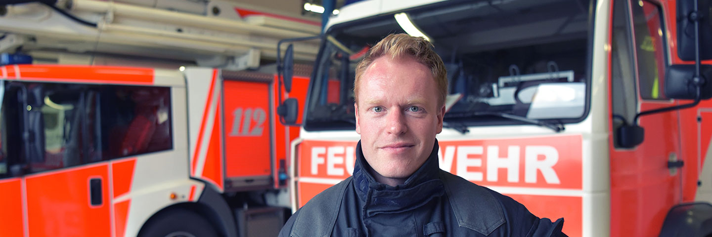 Feuerwehrmann in Feuerwehruniform vor zwei Einsatzfahrzeugen in einer Feuerwehrhalle.