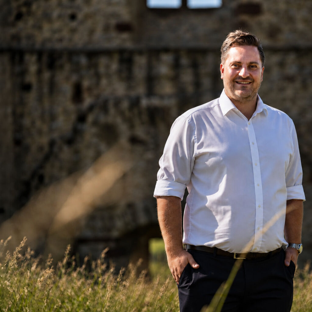Dr. Johannes Zierden im weißen Hemd vor einer alten Burgmauer auf einer Wiese.