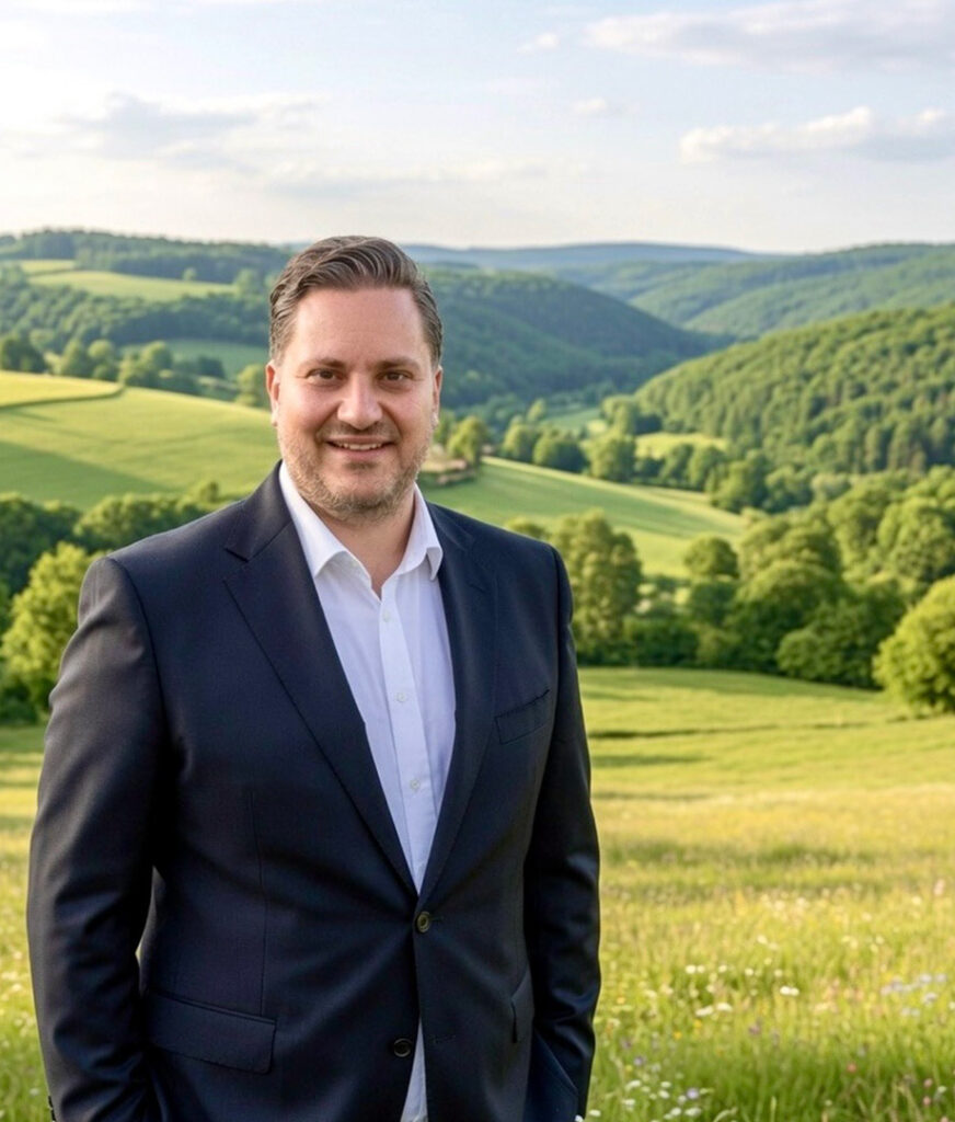 Dr. Johannes Zierden im dunklen Anzug vor einer grünen Hügel- und Wiesenlandschaft der Eifel.