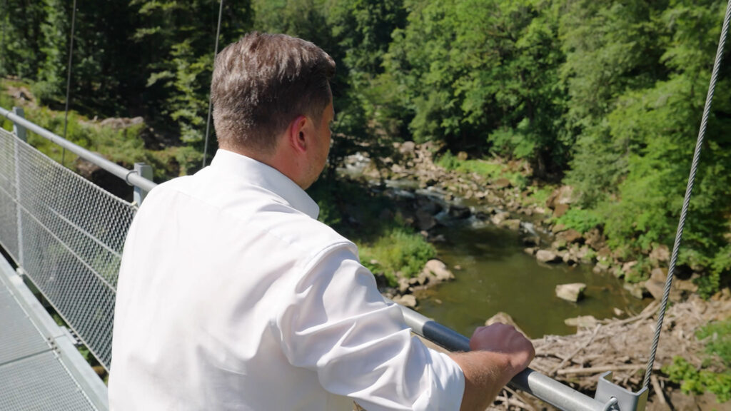 Dr. Johannes Zierden steht auf einer Hängebrücke und blickt auf einen felsigen Bachlauf im Eifelwald.