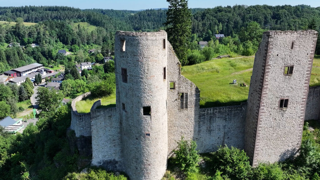 Luftaufnahme der Burgruine Neuerburg mit rundem und eckigem Turm über dem bewaldeten Tal.