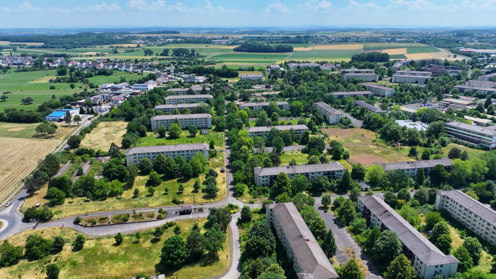 Luftaufnahme einer parkähnlichen Wohnsiedlung mit langen, gleichförmigen Gebäudezeilen zwischen Feldern in der Eifel.